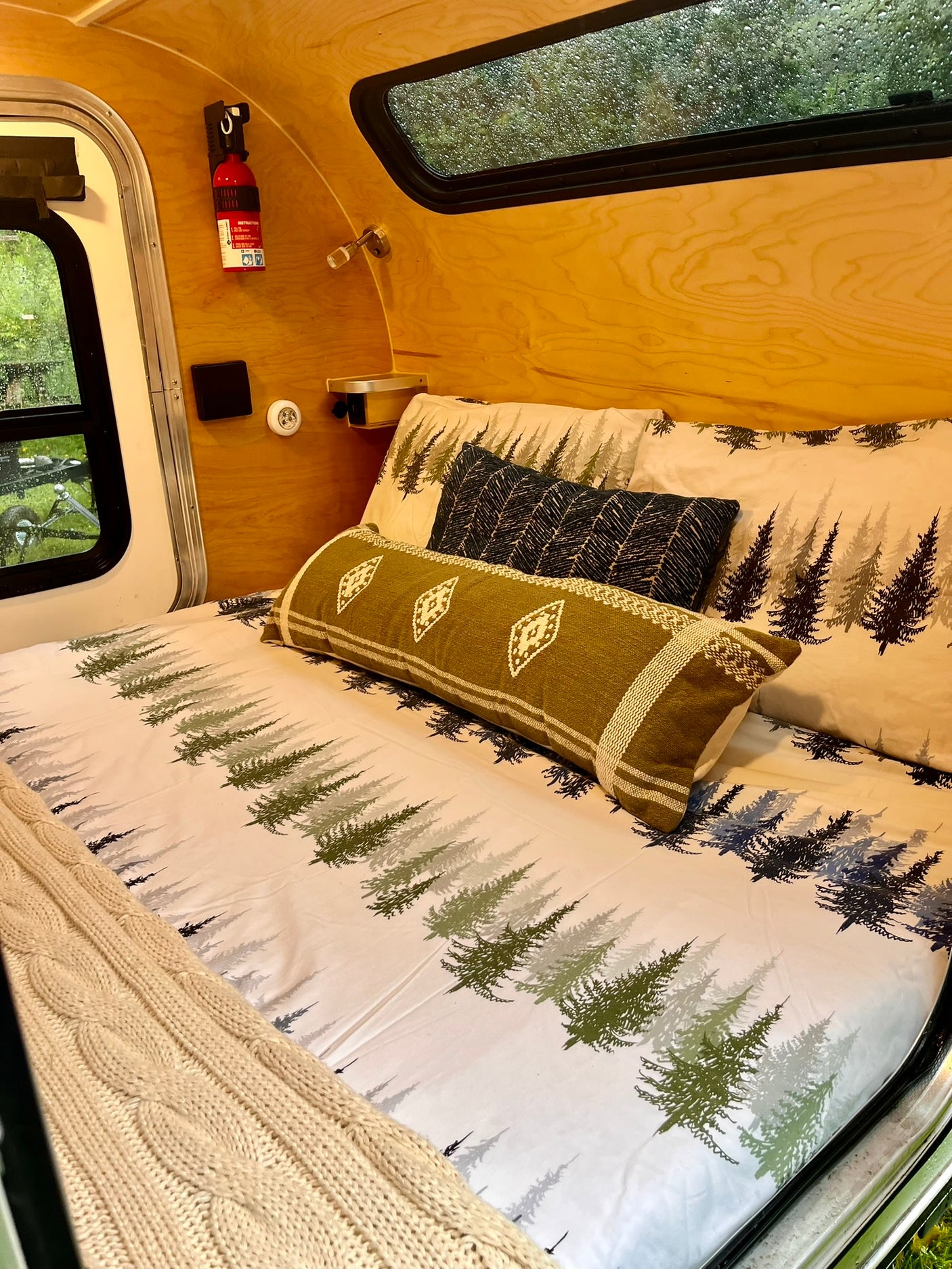 Cozy RV rental interior features a bed with white forest-print bedding, decorative pillows, and a knit blanket. Wood-paneled walls.