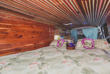 Step into this unique RV rental: a cozy bed with ukulele & leis, surrounded by custom wood walls and ceiling.
