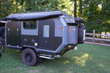 Discover this compact overland trailer, a perfect rv rental for adventures! Gray & black design, off-road tires, parked on grass.