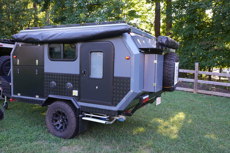 Discover this compact overland trailer, a perfect rv rental for adventures! Gray & black design, off-road tires, parked on grass.