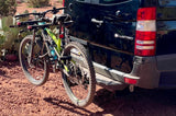 Two mountain bikes are secured on an RV rental van's rear rack, parked on red desert ground, ready for adventure.
