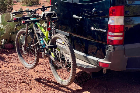 Two mountain bikes are secured on an RV rental van's rear rack, parked on red desert ground, ready for adventure.