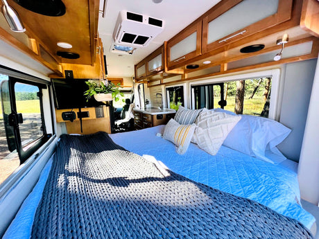 Bright RV rental interior with a cozy bed, TV, and kitchen. Large windows offer scenic outdoor views, ideal for comfortable travel.