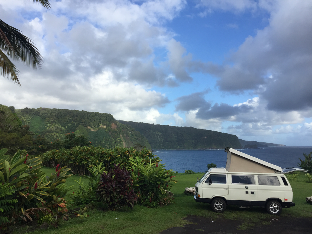 White pop-top RV rental camper van parked on a grassy cliff overlooking a blue ocean & green mountains.