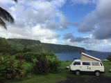White pop-top RV rental camper van parked on a grassy cliff overlooking a blue ocean & green mountains.