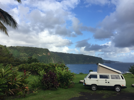 White pop-top RV rental camper van parked on a grassy cliff overlooking a blue ocean & green mountains.