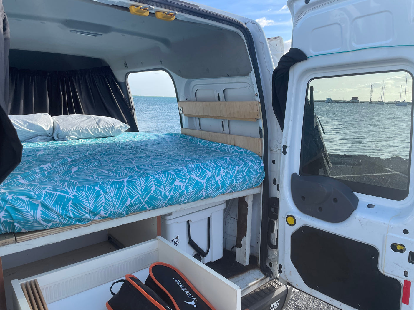 RV rental camper van interior. Features a comfy bed, storage, & ocean view from open doors.
