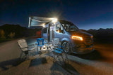 RV rental campout at night: a blue camper van with bright awning lights, headlights, camp chairs, and a lantern-lit table.