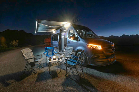 RV rental campout at night: a blue camper van with bright awning lights, headlights, camp chairs, and a lantern-lit table.