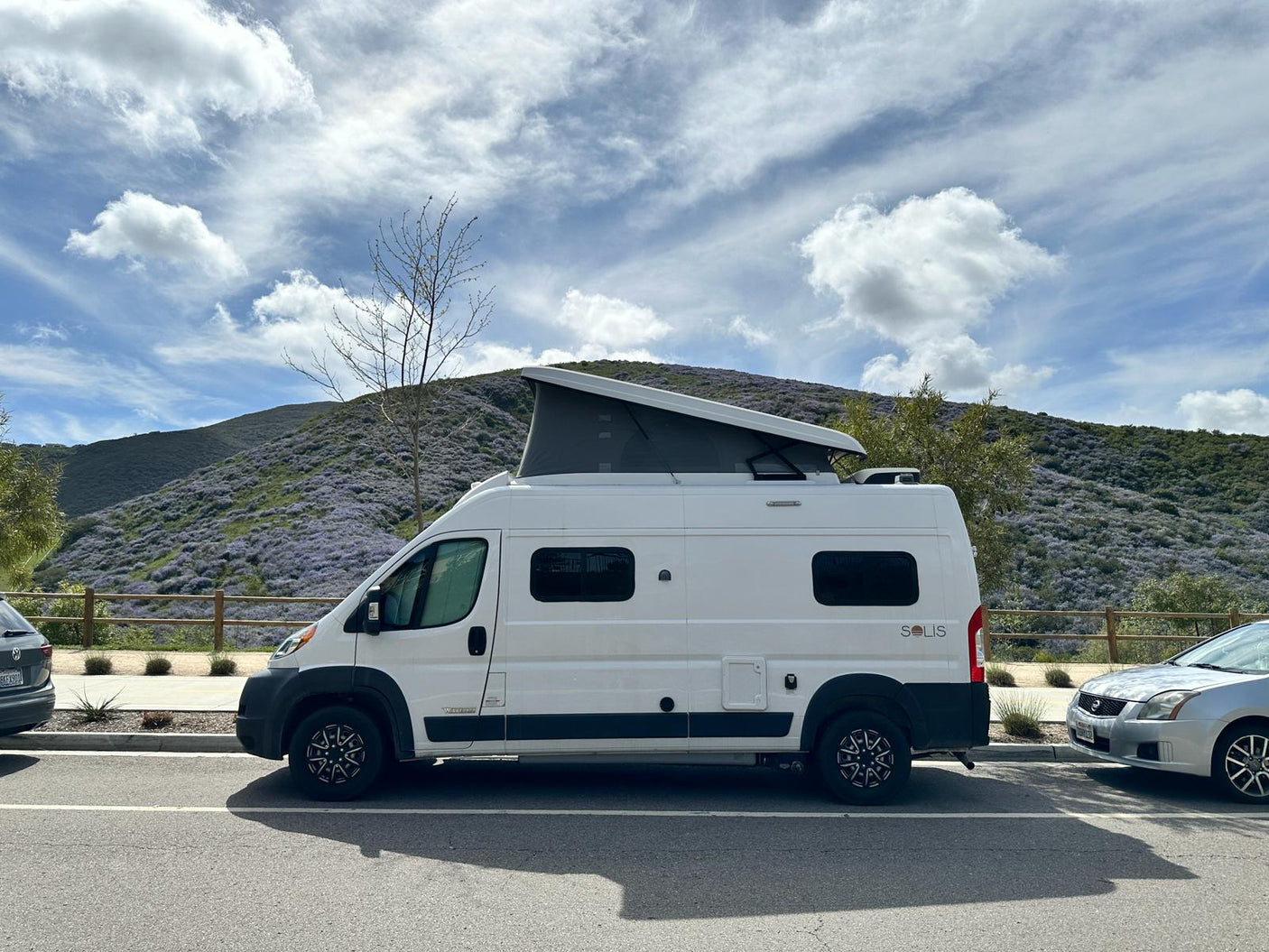 White Winnebago Solis camper van with pop-top, ideal for rv rental adventures, parked by a hill covered in vibrant purple flowers.