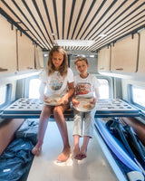 Two children enjoy sandwiches inside an RV rental. Visible are the raised bed frame, overhead cabinets, and floor storage.