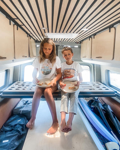 Two children enjoy sandwiches inside an RV rental. Visible are the raised bed frame, overhead cabinets, and floor storage.