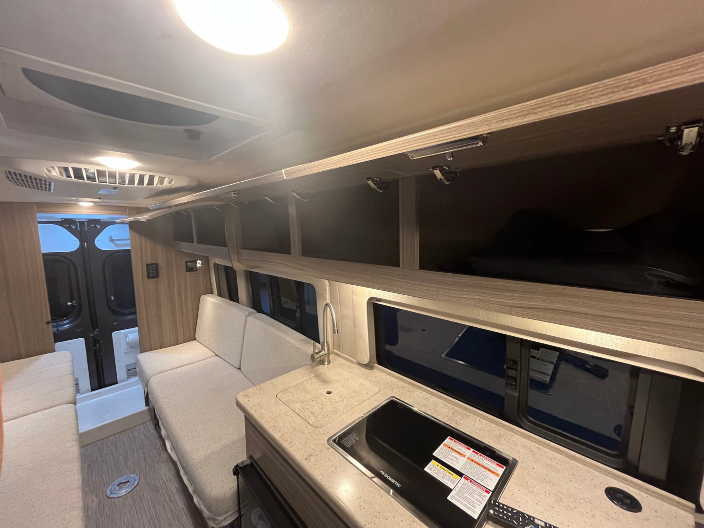 Bright RV rental interior showing a kitchen with sink and Dometic cooktop, sofa seating, and overhead storage.