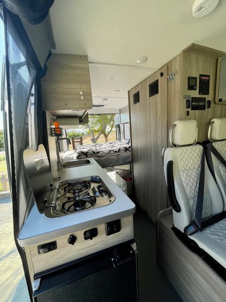 Bright interior of a modern rv rental, featuring a compact kitchen, cozy bed, and white passenger seats.