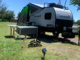 A light-colored Viking RV rental is set up for outdoor living, featuring an awning, grill, and prep table, under a clear sky.