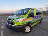 Vibrant custom-painted Ford Transit van with graffiti art, possibly an rv rental, parked outdoors with mountains and city in the background.