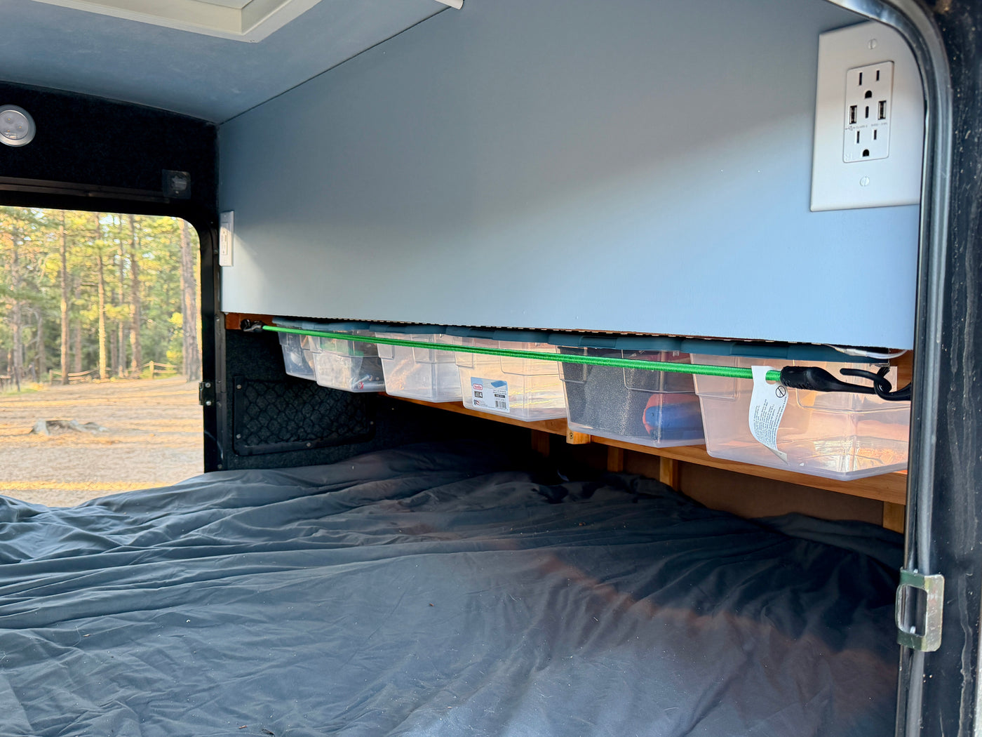 RV rental bedroom with dark bed, overhead storage bins, and power outlet. A green bungee secures bins. Forest outside window. — photo 9