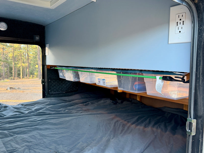RV rental bedroom with dark bed, overhead storage bins, and power outlet. A green bungee secures bins. Forest outside window. — photo 9