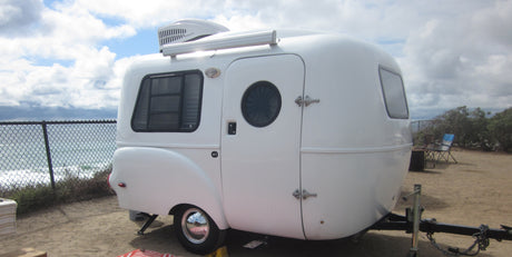Oceanfront RV rental: A sleek white teardrop trailer with an AC unit, parked steps from the ocean for seaside camping.