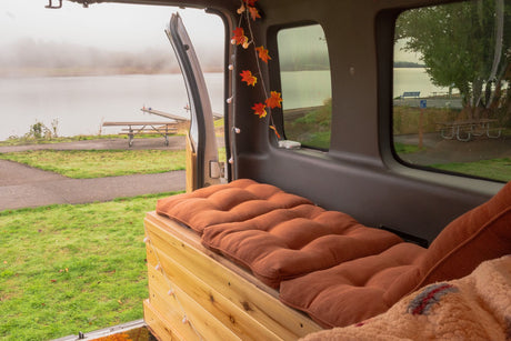 An RV rental's cozy interior, adorned with autumn lights, offers a tranquil view of a misty lake and park.