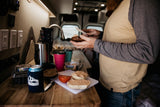RV rental kitchen: A person spreads a meal on bread inside a custom van, with coffee & breakfast items on the counter.