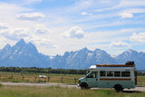 An RV rental bus, light blue & white, faces majestic Grand Teton mountains & a field. Ready for adventure!