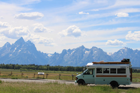 An RV rental bus, light blue & white, faces majestic Grand Teton mountains & a field. Ready for adventure!