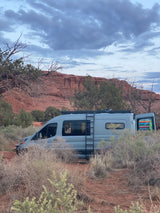 Discover adventure with an **RV rental**! A grey camper van is parked in a desert landscape with red rock formations under a cloudy sky.