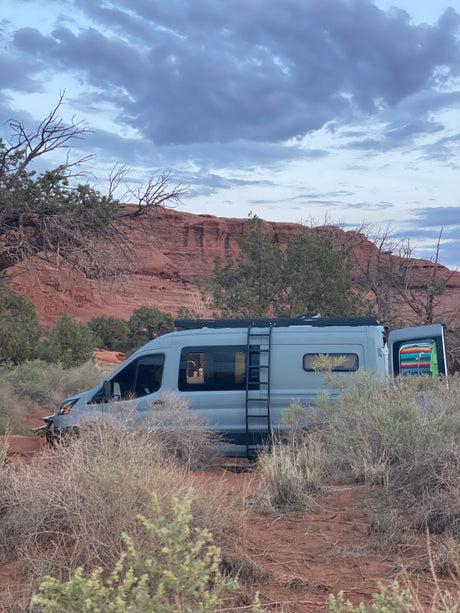 Discover adventure with an **RV rental**! A grey camper van is parked in a desert landscape with red rock formations under a cloudy sky.