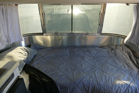 Inside an rv rental bedroom: a large bed with gray bedding, wide windows, and bright sunlight.