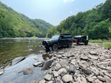 Dark SUV & off-road RV rental parked on a rocky riverbank. Lush green mountains line a clear river, ideal for adventure.
