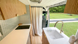 Step into this chic RV rental! Features a compact kitchen with a sink, cooktop, wood cabinets, & outdoor views.