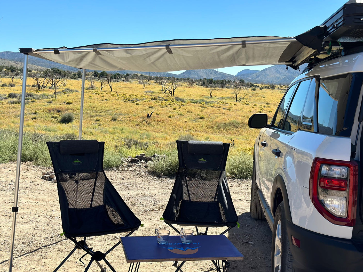 "RV rental" trip setup: A white SUV with an attached awning shades two chairs and a table in a sunny, dry desert landscape.