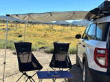 "RV rental" trip setup: A white SUV with an attached awning shades two chairs and a table in a sunny, dry desert landscape.