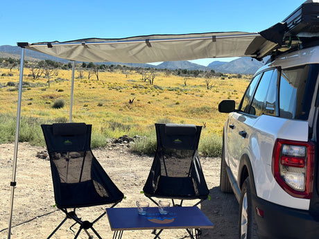 "RV rental" trip setup: A white SUV with an attached awning shades two chairs and a table in a sunny, dry desert landscape.