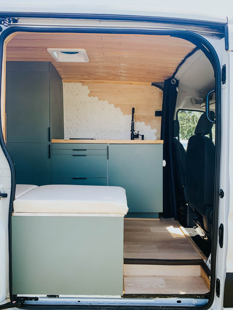 Modern camper van interior for RV rental: custom kitchen, bed, wood ceiling, & green cabinets.