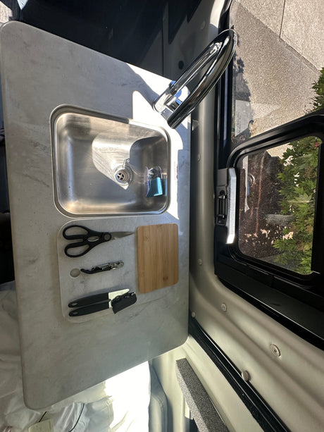rv rental: functional kitchen counter featuring a stainless steel sink, faucet, cutting board, and kitchen tools by a window.