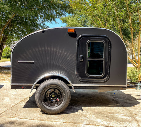 Side view of a sleek black teardrop camper, great for an rv rental. Parked on concrete under sunny trees.