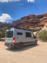 A gray RV rental van, parked on a dirt lot, faces stunning red rock cliffs under a clear blue sky. Ideal for wild adventures.