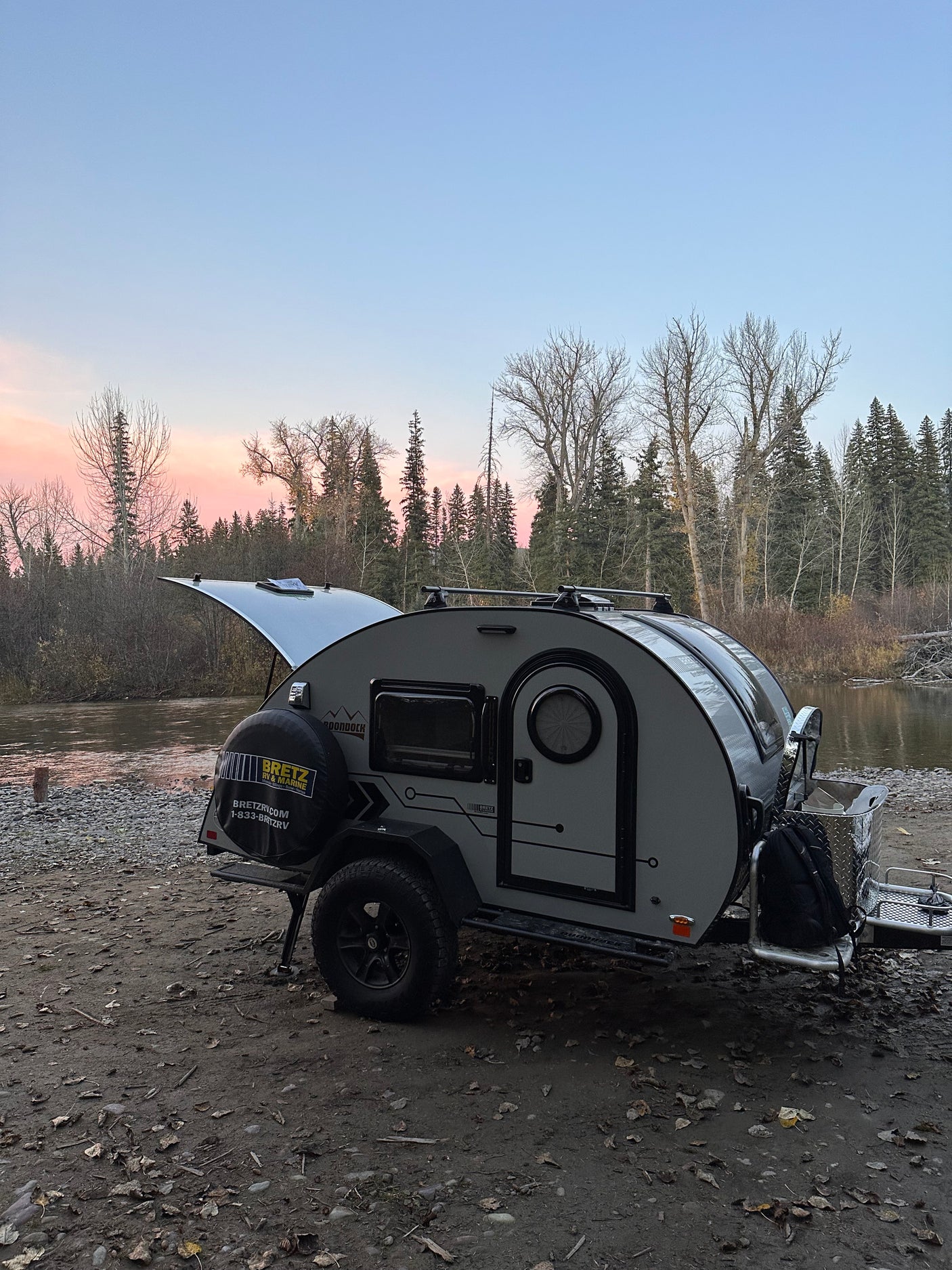 Light grey RV rental teardrop camper parked by a river at pink sunset, rear hatch open for adventure.