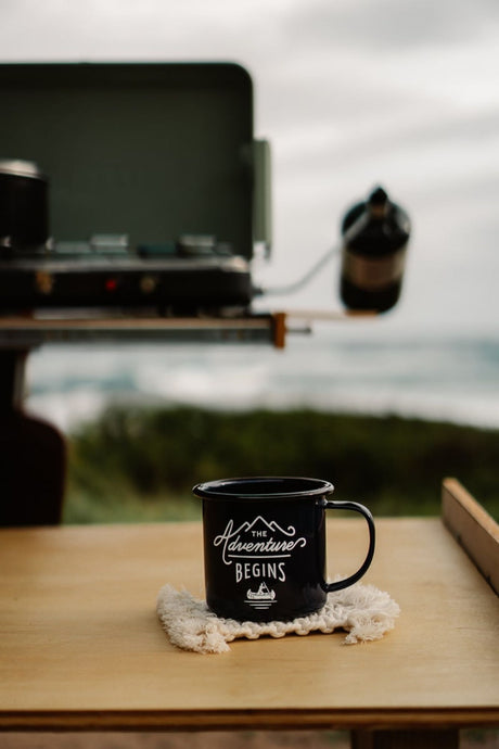 Enjoy morning coffee from your RV rental. A black enamel "Adventure Begins" mug on a wooden table, with a camp stove and ocean view in the blurred background.