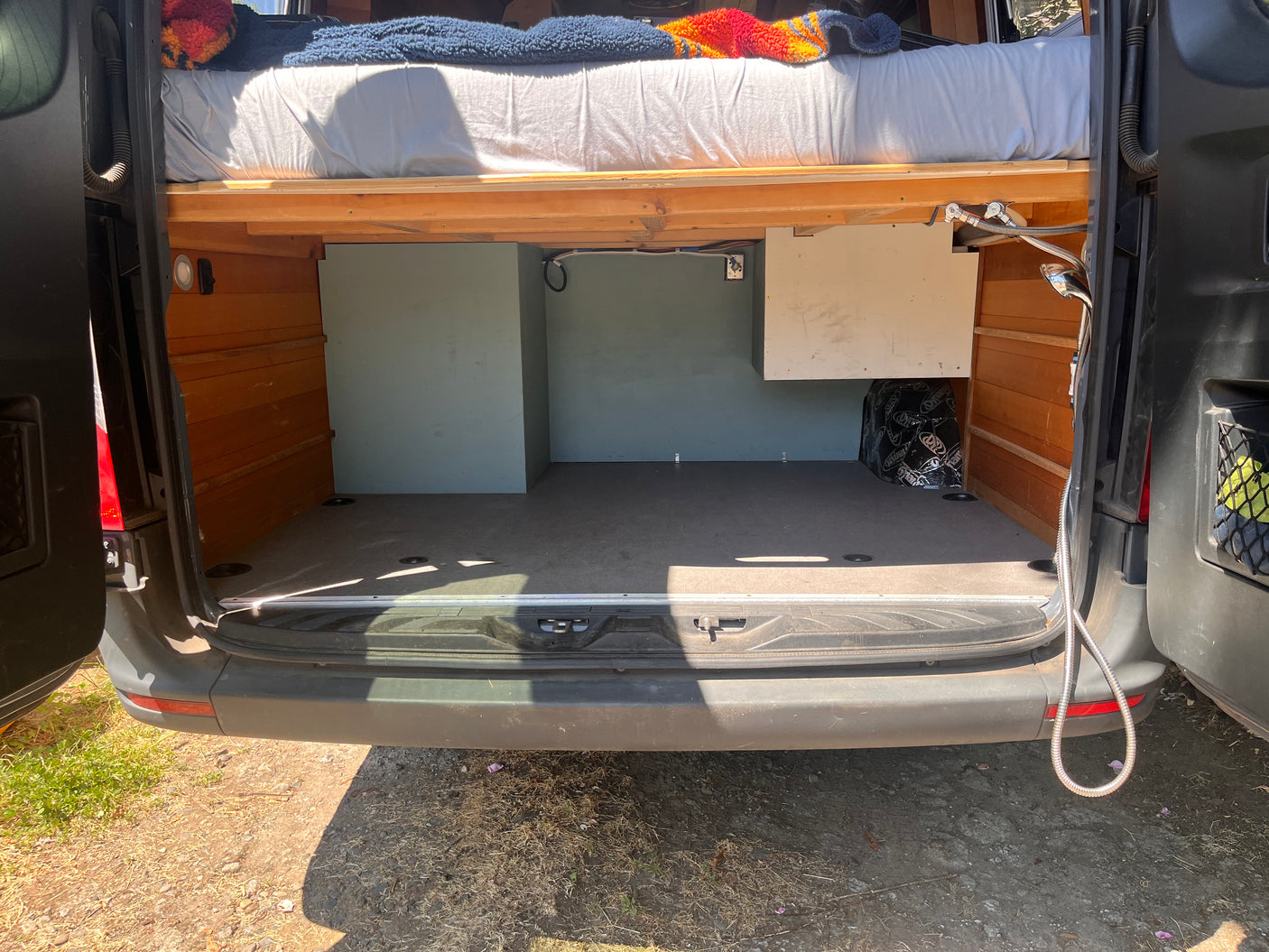 Rear view of an RV rental campervan interior with raised bed, ample cargo space, storage, and outdoor shower access.