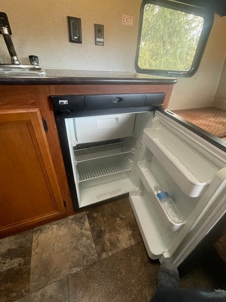 Inside an RV rental: a compact kitchen area featuring an open refrigerator, sink, and counter. An "EXIT" sign is above. Ready for travel!