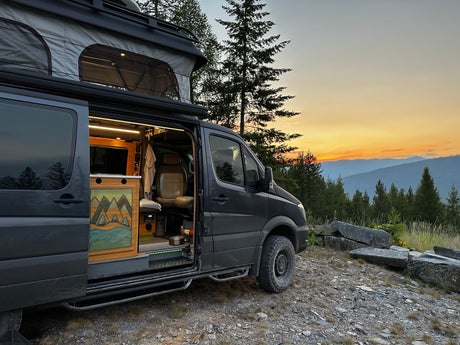 This pop-top camper van RV rental is parked with its door open, showing a warm interior against a stunning mountain sunset.