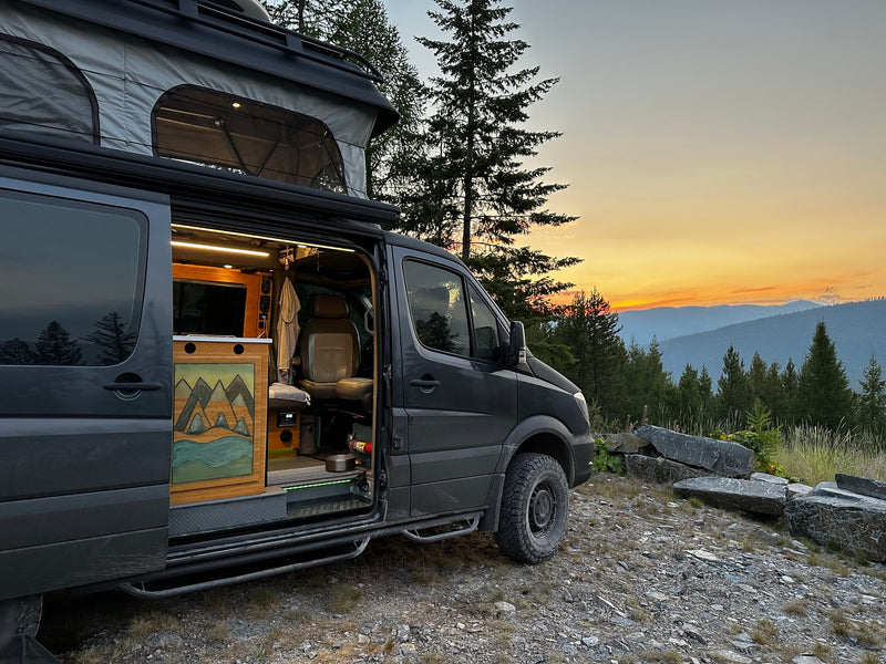 This pop-top camper van RV rental is parked with its door open, showing a warm interior against a stunning mountain sunset. — photo 3