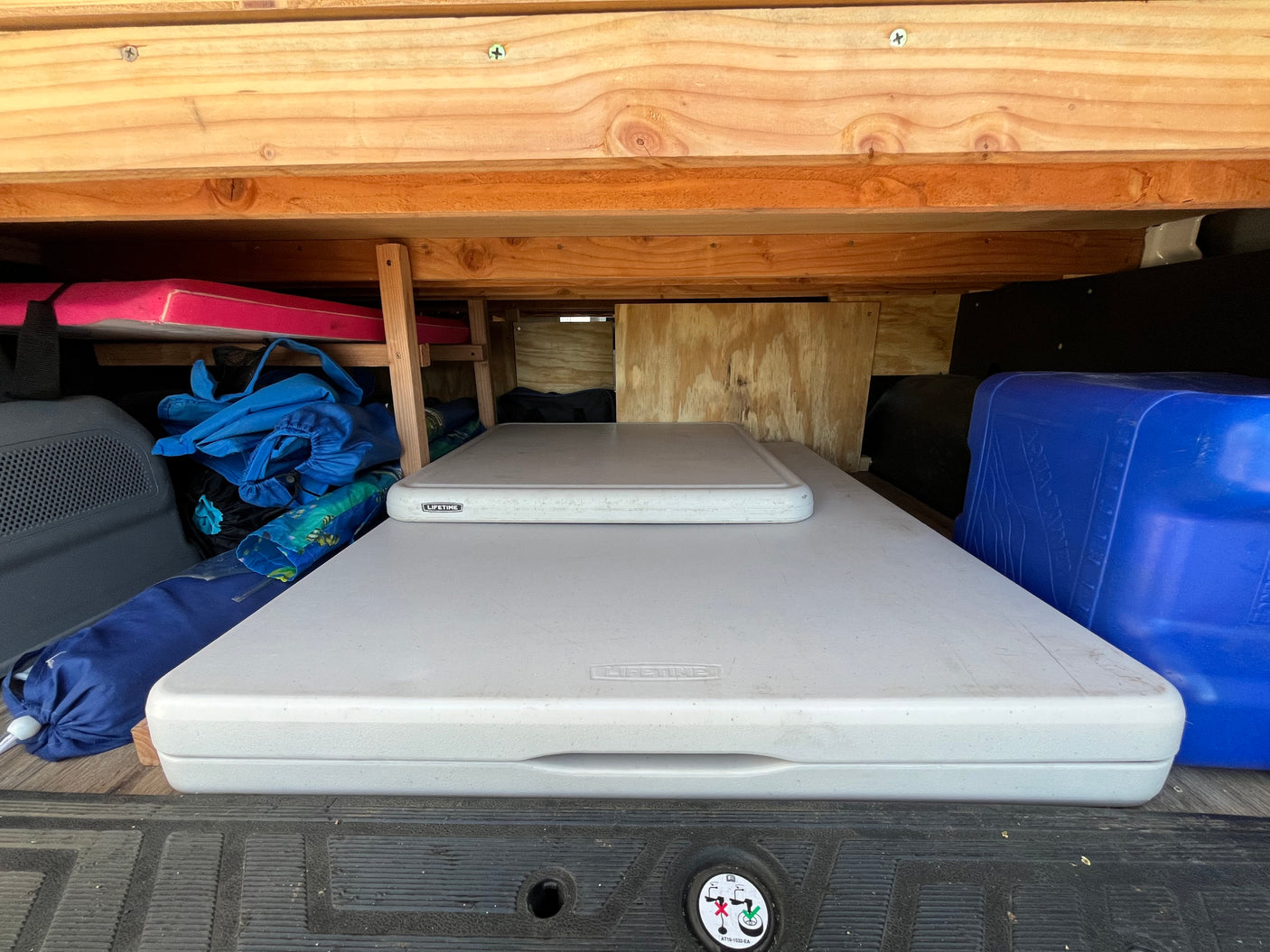 RV rental organized storage: Two white Lifetime tables, blue water jug, bags & pink mattress under wooden shelves in a van.