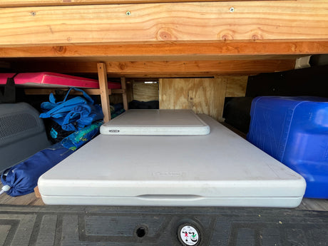 RV rental organized storage: Two white Lifetime tables, blue water jug, bags & pink mattress under wooden shelves in a van.
