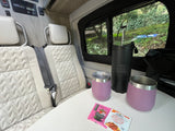 Relaxing RV rental interior: Seats, table with tea/mugs. Solar power setup on the wall. Lush greenery outside window.