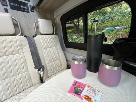 Relaxing RV rental interior: Seats, table with tea/mugs. Solar power setup on the wall. Lush greenery outside window.