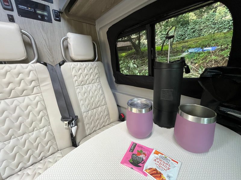 Relaxing RV rental interior: Seats, table with tea/mugs. Solar power setup on the wall. Lush greenery outside window. — photo 8